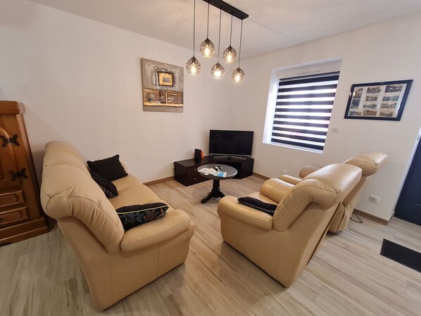 Un salon avec deux canapés beige, une table basse, un téléviseur et une fenêtre avec des stores.