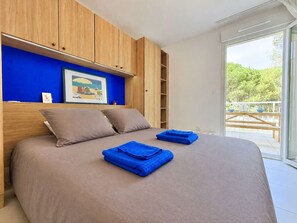 A bedroom with a bed, blue towels, wooden cabinets, a framed picture, and a view of greenery outside. - Apartment with AC and parking - 4 guests (Le Lavandou)