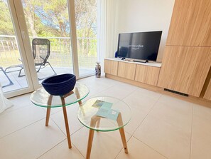 Ein modernes Wohnzimmer mit einem Glastisch, einer blauen Schale, einem Fernseher auf einer Holzkommode und Blick auf einen Balkon.
