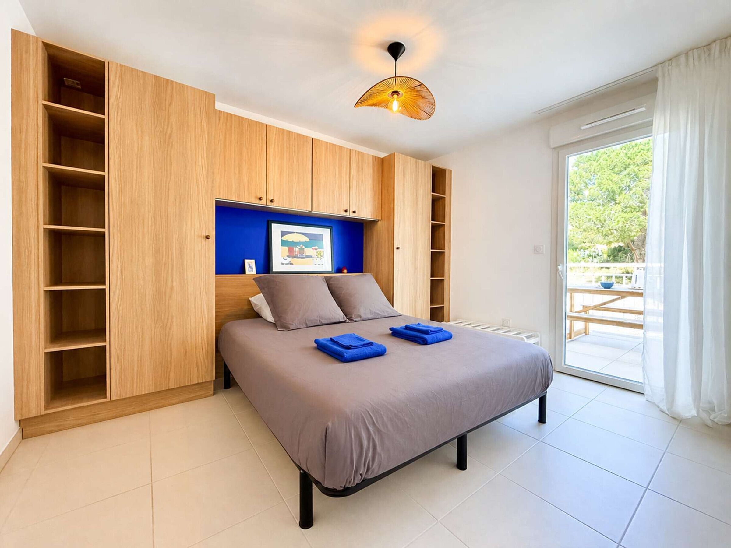 Ein Schlafzimmer mit einem Bett, einer blauen Wand, einem Holzschrank und Blick ins Freie.