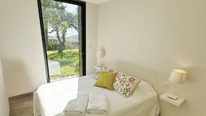 Une chambre à coucher avec un lit, des oreillers, une table de chevet, une lampe et une vue sur la verdure par la fenêtre.