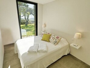 Ein Schlafzimmer mit Bett, Kissen, einem Nachttisch, einer Lampe und Blick auf Grün durch ein Fenster.