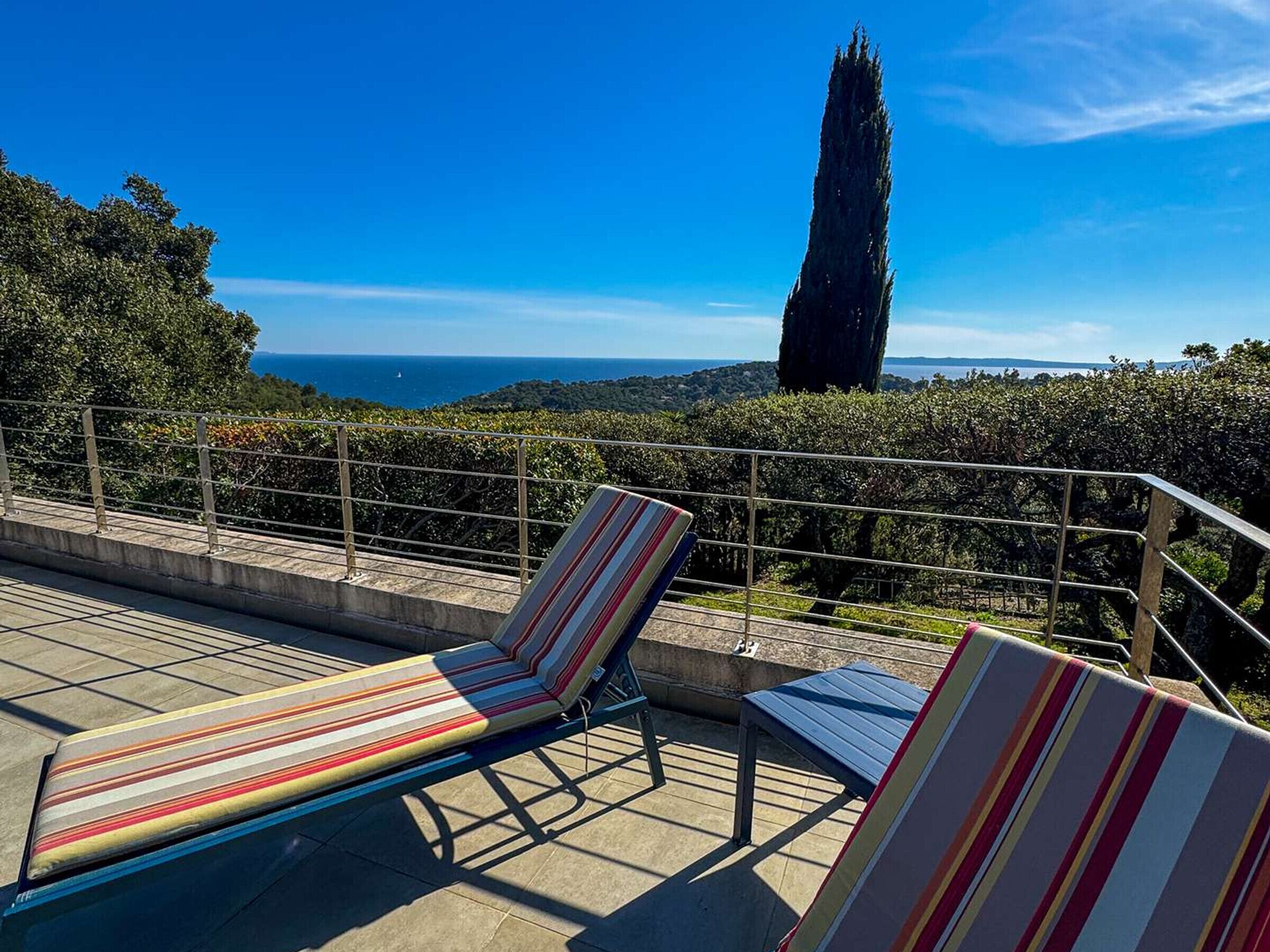 Una terraza con dos sillones a rayas, una barandilla de metal y vistas al mar y a un árbol alto.
