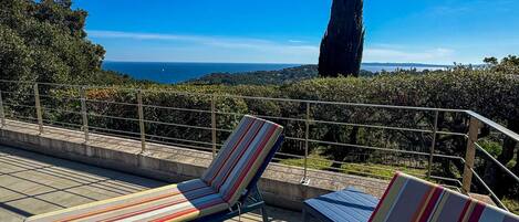 Una terraza con dos sillones a rayas, una barandilla de metal y vistas al mar y a un árbol alto.