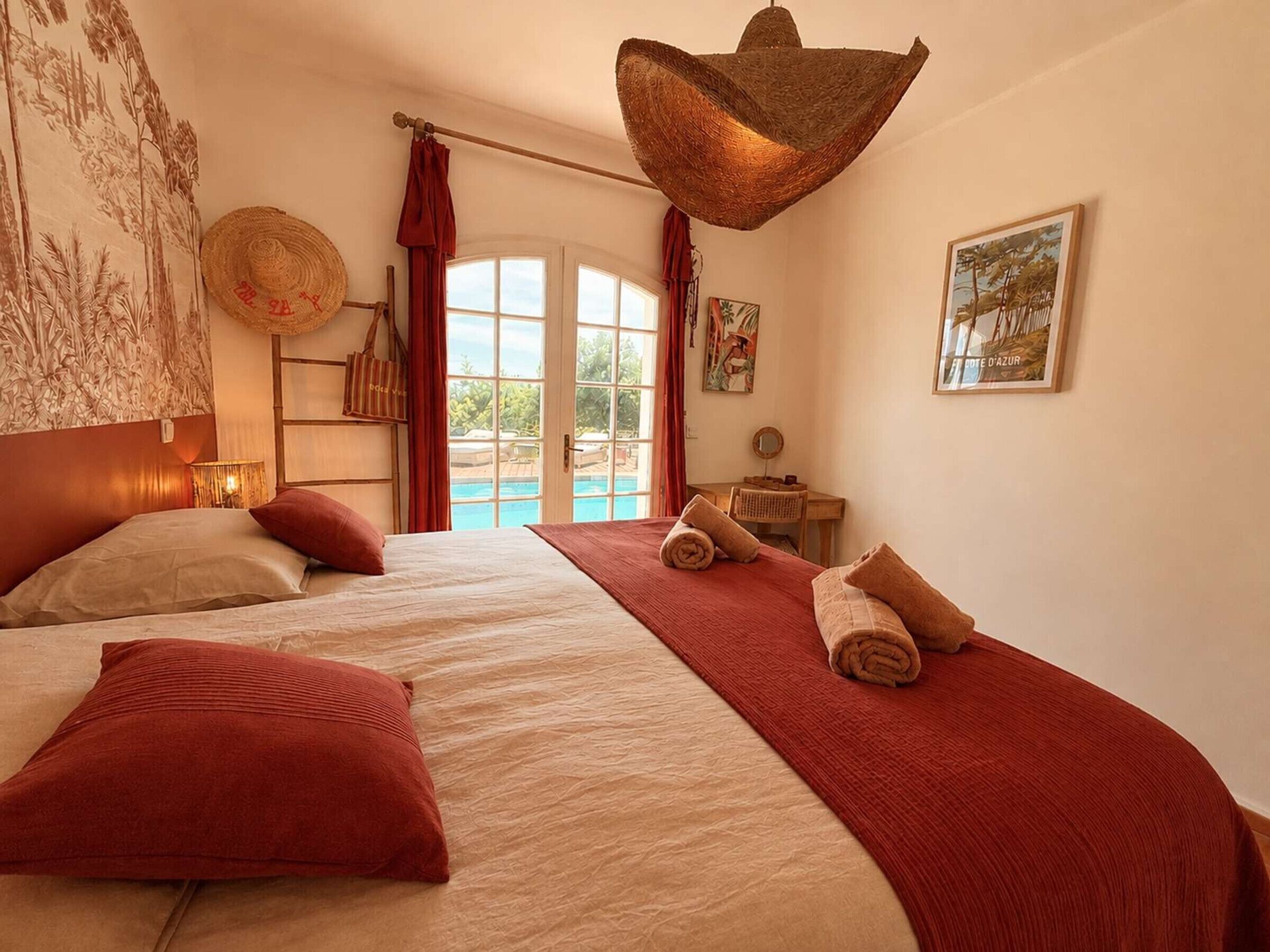 A hotel room with a large bed, a desk, a chair, a hanging light fixture, and a view of a pool.