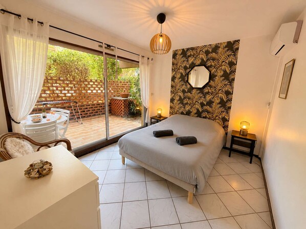Een slaapkamer met een bed, een bureau en een stoel. Er is een glazen schuifdeur die leidt naar een terras met een tafel en stoelen.