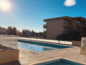 A swimming pool with a stone deck, surrounded by a white metal fence, and a multi-story building in the background. - Ground floor apartment, AC - 3 guests (Bormes-les-Mimosas)
