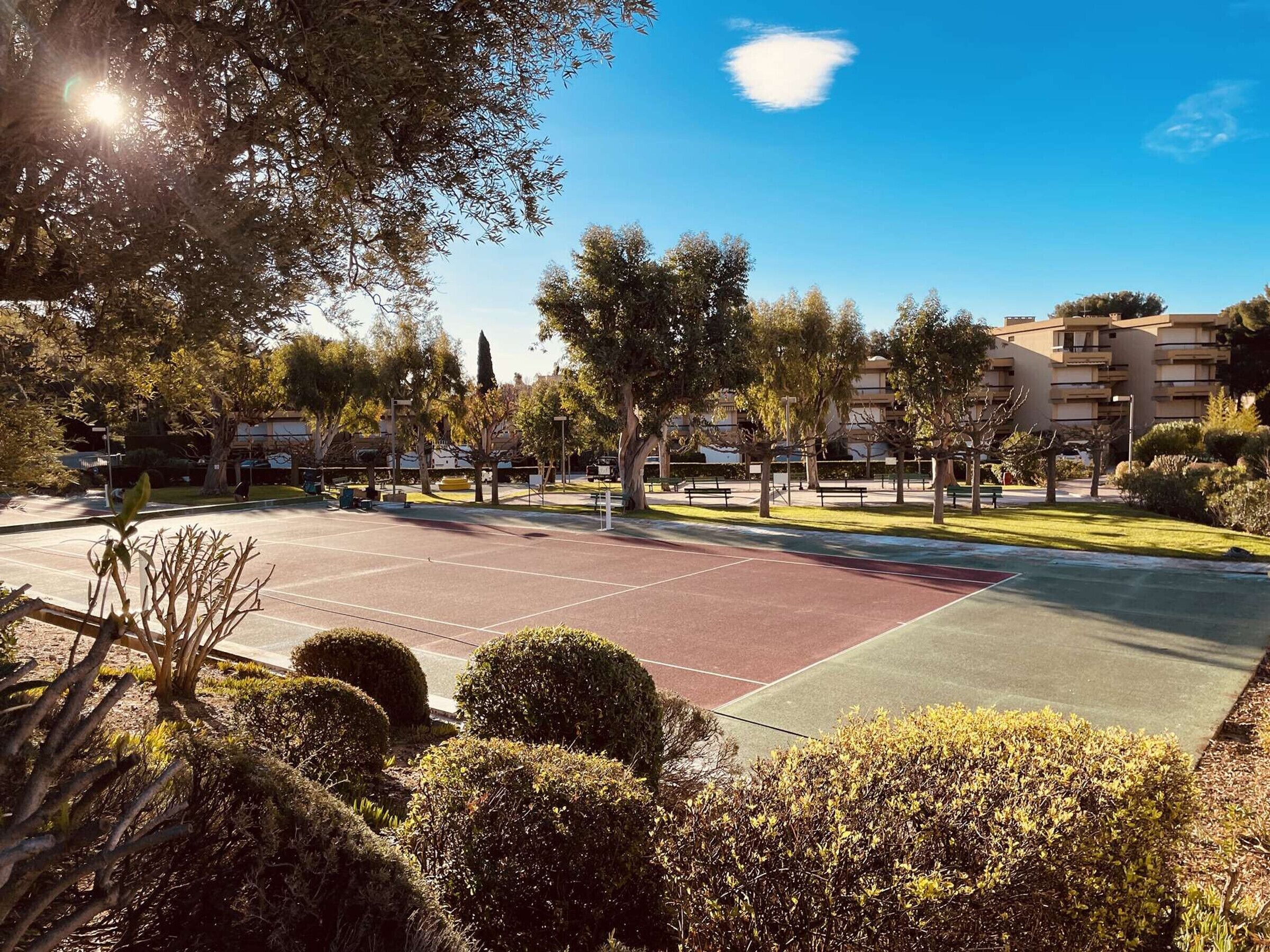 A tennis court surrounded by well-maintained bushes and trees, with a clear sky above.