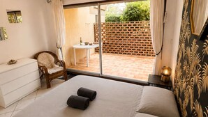 Ein Schlafzimmer mit einem Bett, einem Sessel, einem kleinen Tisch mit Flasche und Schale, einer Pendelleuchte und Blick auf eine Terrasse mit Ziegelmauer und Grünpflanzen.