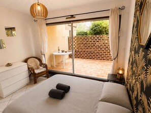 A bedroom with a bed, a chair, a small table with a bottle and bowl, a hanging light fixture, and a view of a patio with a brick wall and greenery. - Ground floor apartment, AC - 3 guests (Bormes-les-Mimosas)