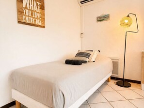 A minimalist bedroom with a single bed, a wall-mounted air conditioner, a floor lamp, and a motivational poster. - Ground floor apartment, AC - 3 guests (Bormes-les-Mimosas)