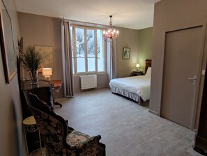 A hotel room with a bed, a desk with a lamp, a chair, a window with curtains, and a chandelier. - Central T2 with garden, ideal - ? guests (La Roche-Posay)
