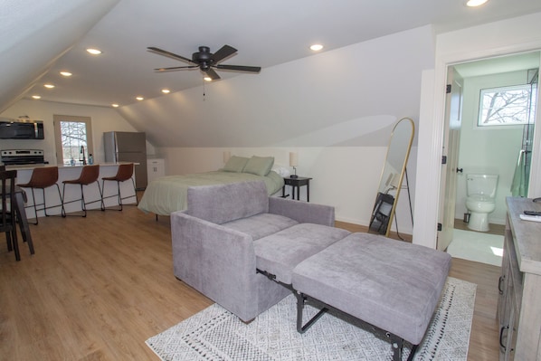 A compact, modern loft bedroom with a ceiling fan, a bed, a small desk, and a kitchenette.