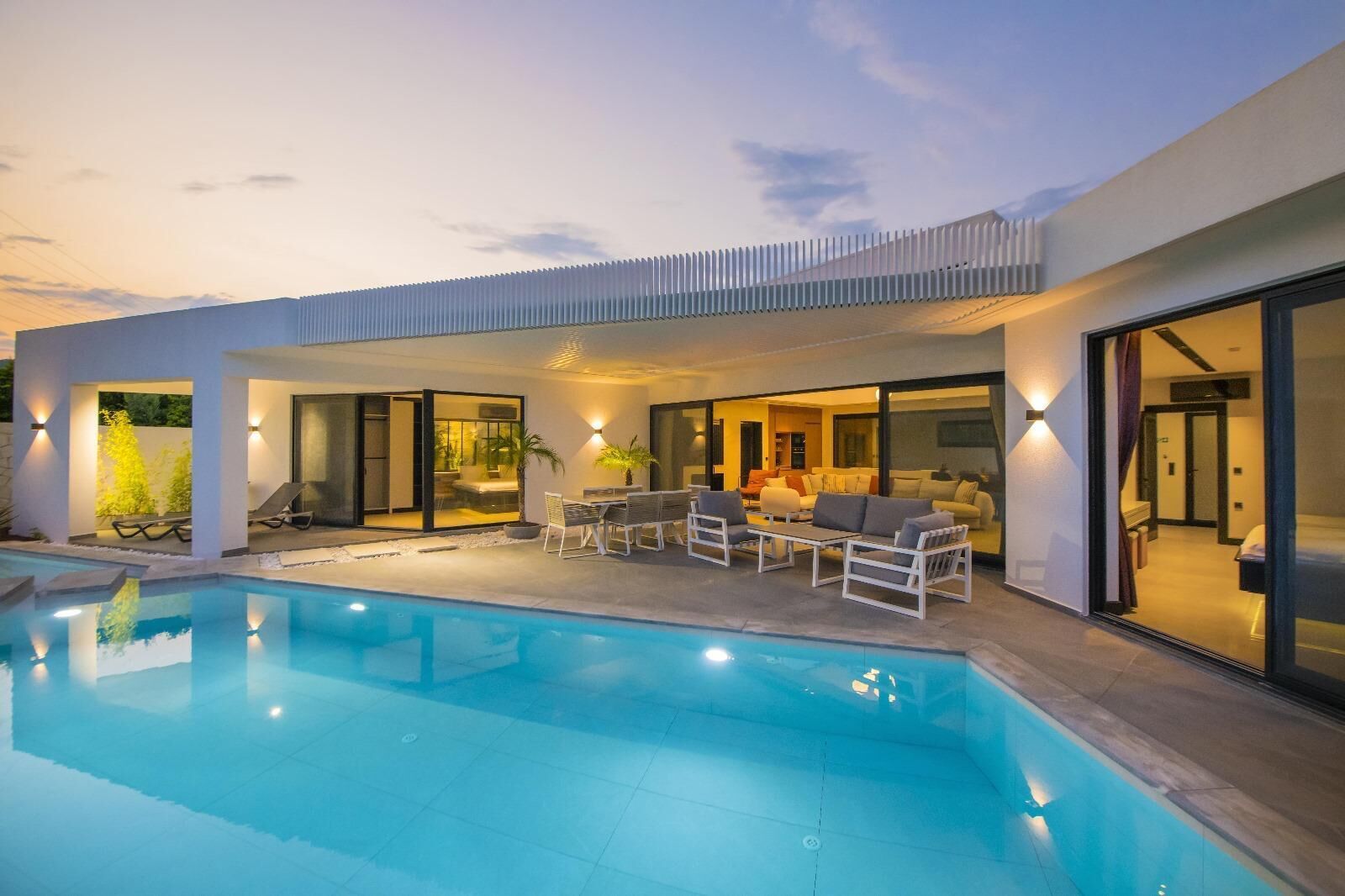 Une maison moderne dotée d’une piscine, d’un espace salon extérieur et de grandes portes vitrées donnant sur l’intérieur.