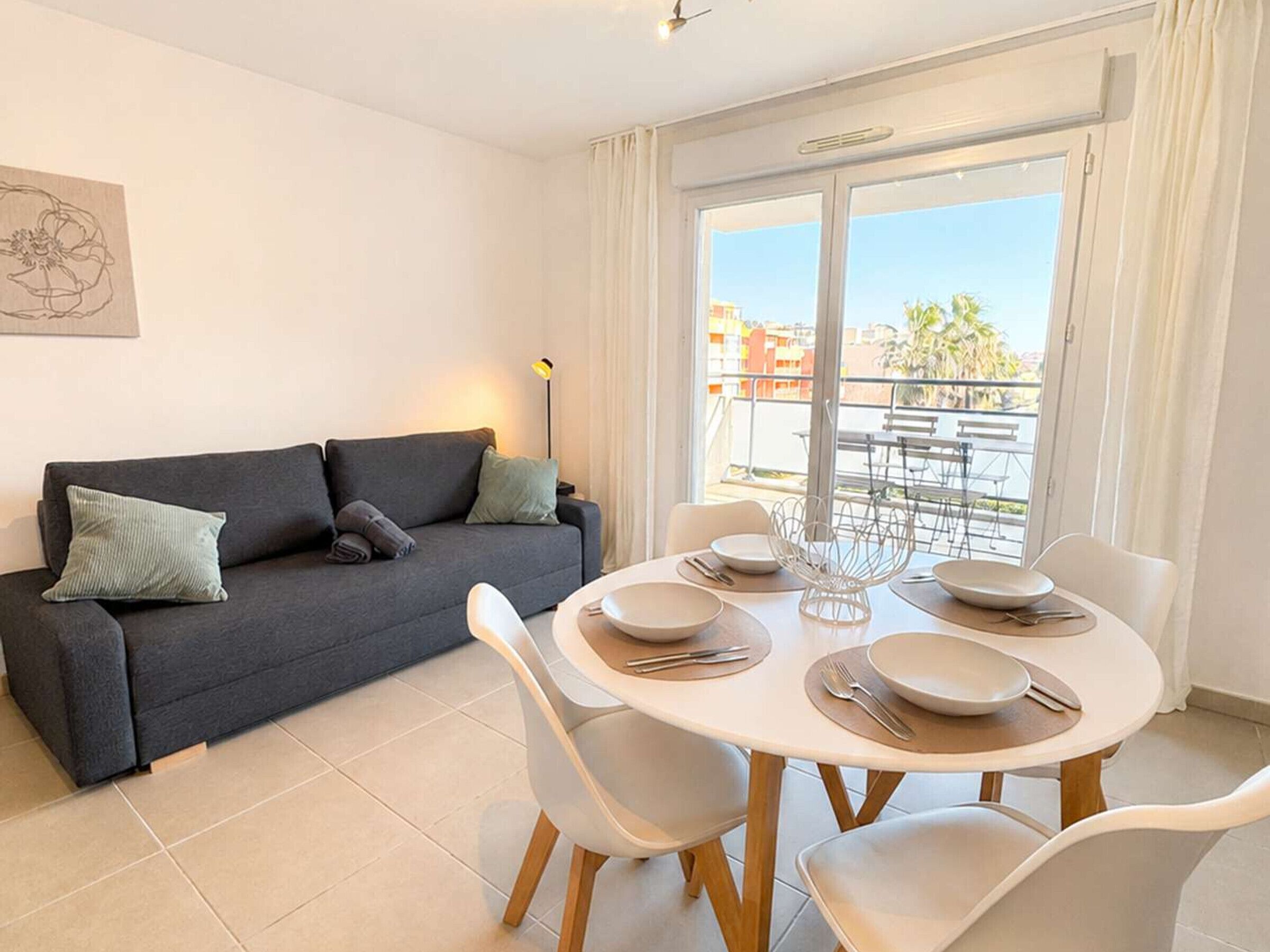 A modern living room with a sofa, dining table, and chairs. There is a balcony with a view of buildings and palm trees.