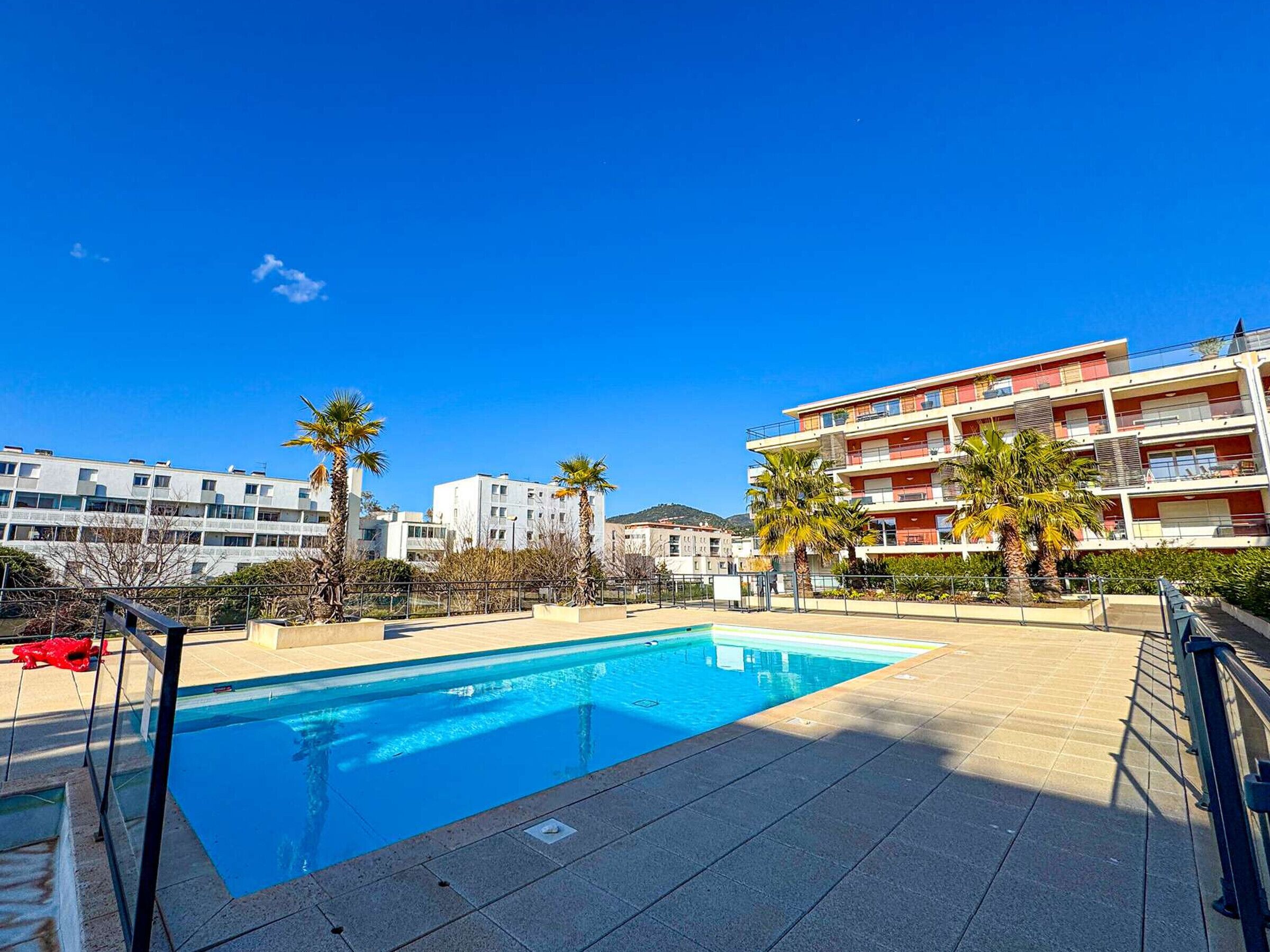 A swimming pool with a clear blue sky, surrounded by palm trees and modern buildings.