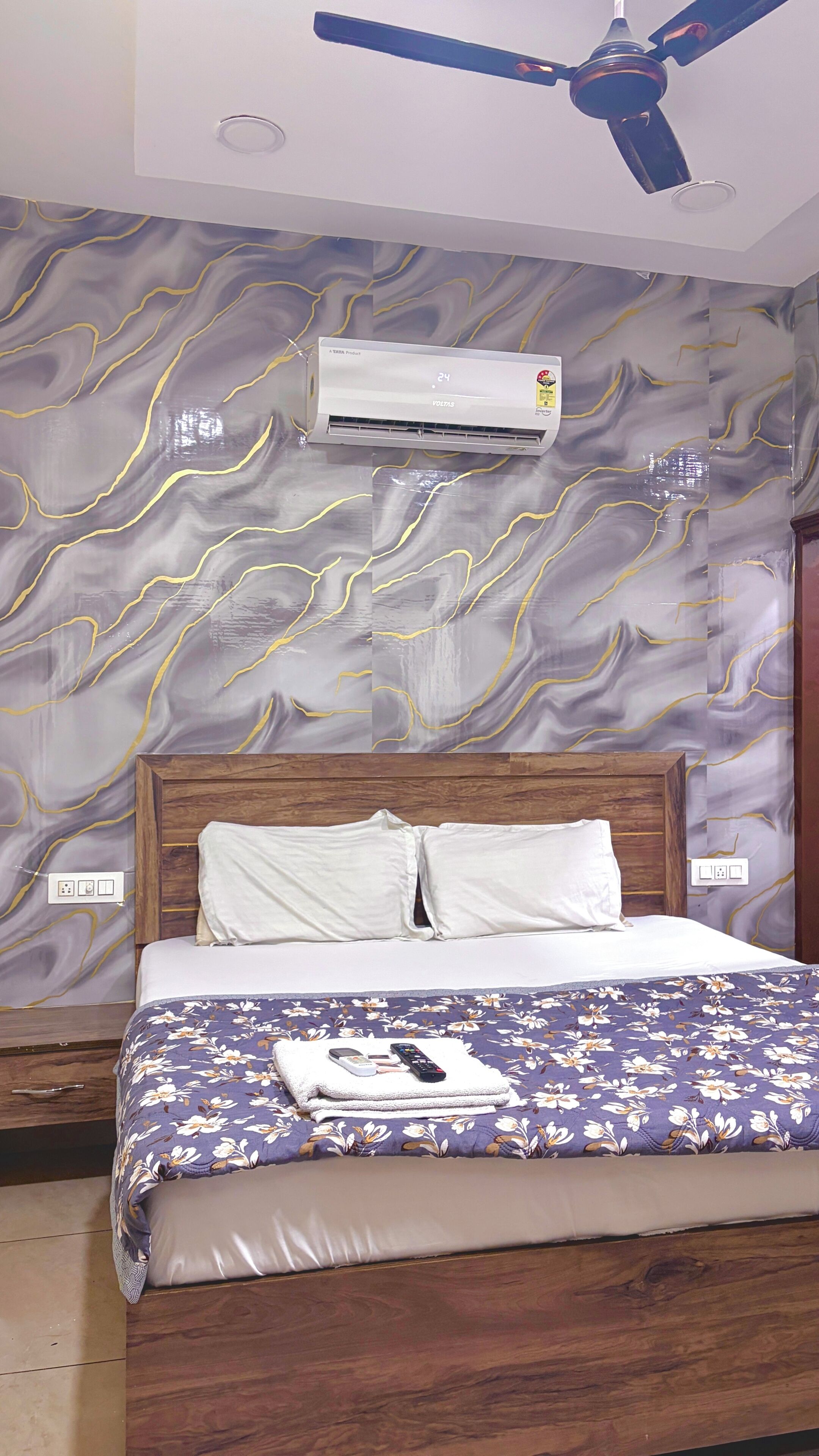 A hotel room with a bed, a ceiling fan, an air conditioning unit, and a wall with a marble-like pattern.