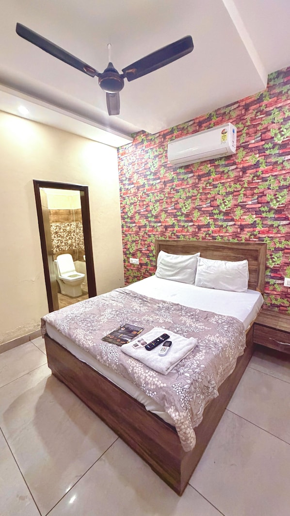 Habitación de hotel con cama, ventilador de techo, aire acondicionado, espejo y baño visible a través de una puerta abierta.