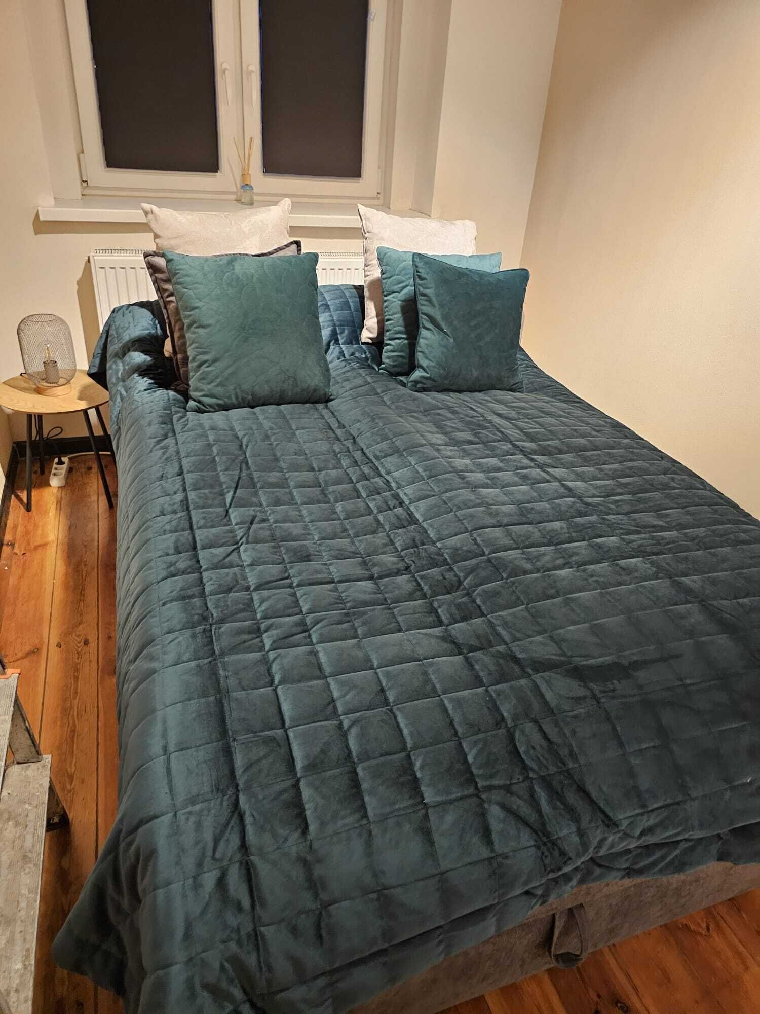 A bed with a dark blue quilt, two pillows, a small bedside table with a lamp, and a window with blinds.