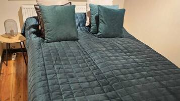 A bed with a dark blue quilt, two pillows, a small bedside table with a lamp, and a window with blinds.