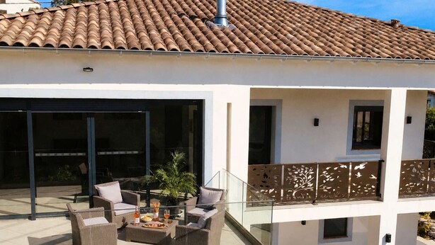 Uma casa moderna com telhado de telhas, varanda coberta com mobiliário externo e guarda-corpo de vidro.