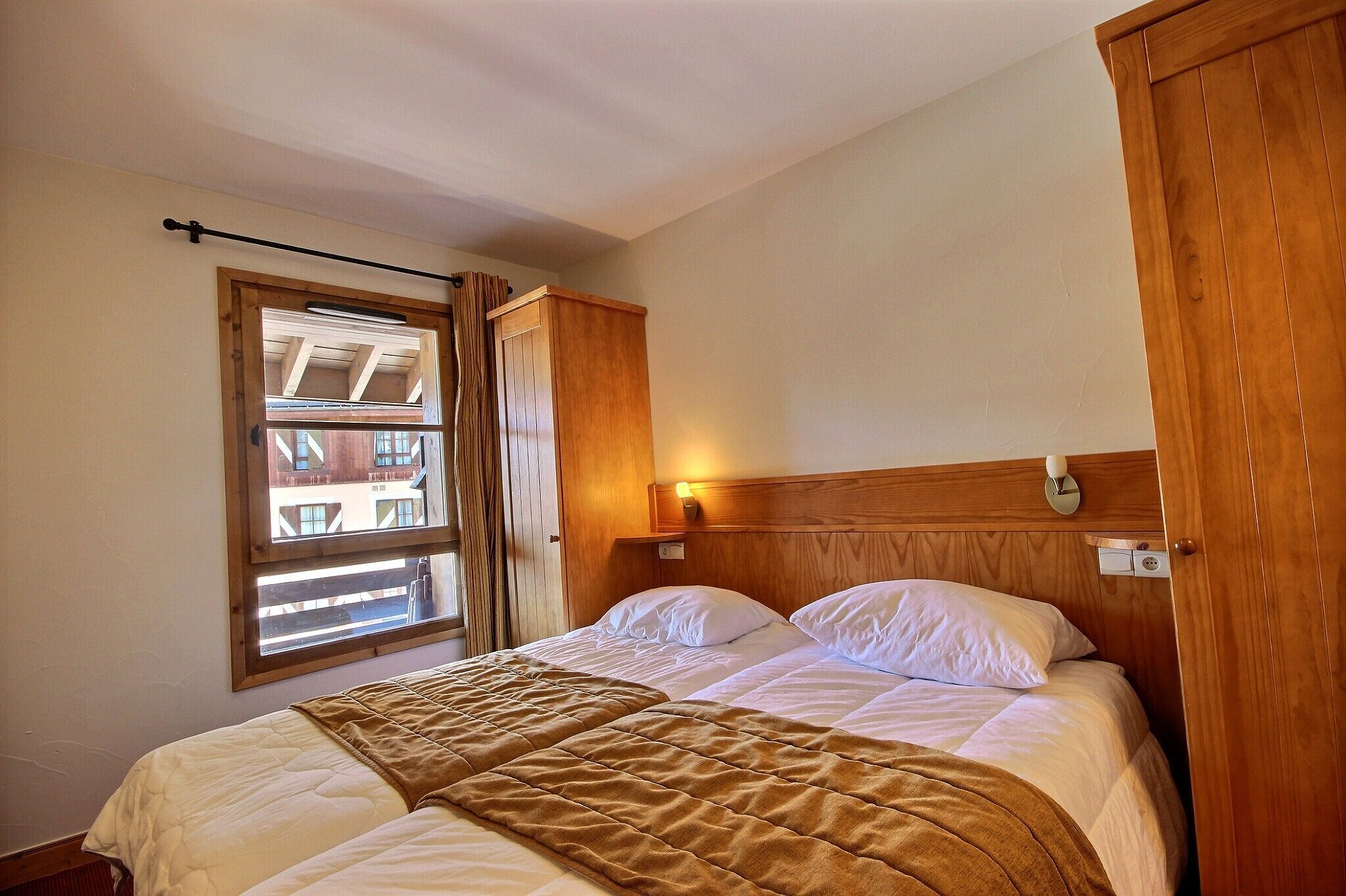 A bedroom with a bed, a window, a wooden wardrobe, and a wall-mounted light fixture.