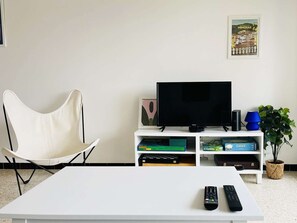 A living room with a white TV stand, a flat-screen TV, a white chair, a coffee table, and a remote control. - Renovated apartment with parking - 6 guests (tennis view) (Bormes-les-Mimosas)