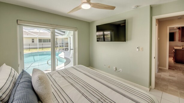 A bedroom with a large bed, a ceiling fan, a TV mounted on the wall, and a sliding glass door leading to a pool area. - Casa Zoe — Sunset Canal Retreat in SE Cape Coral with huge heated pool and spa, bikes, fishing poles, and dartboard (Cape Coral)