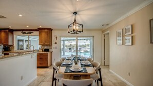 A dining area with a table set for four, a kitchen in the background, and a view of a swimming pool through the window. - Casa Zoe — Sunset Canal Retreat in SE Cape Coral with huge heated pool and spa, bikes, fishing poles, and dartboard (Cape Coral)