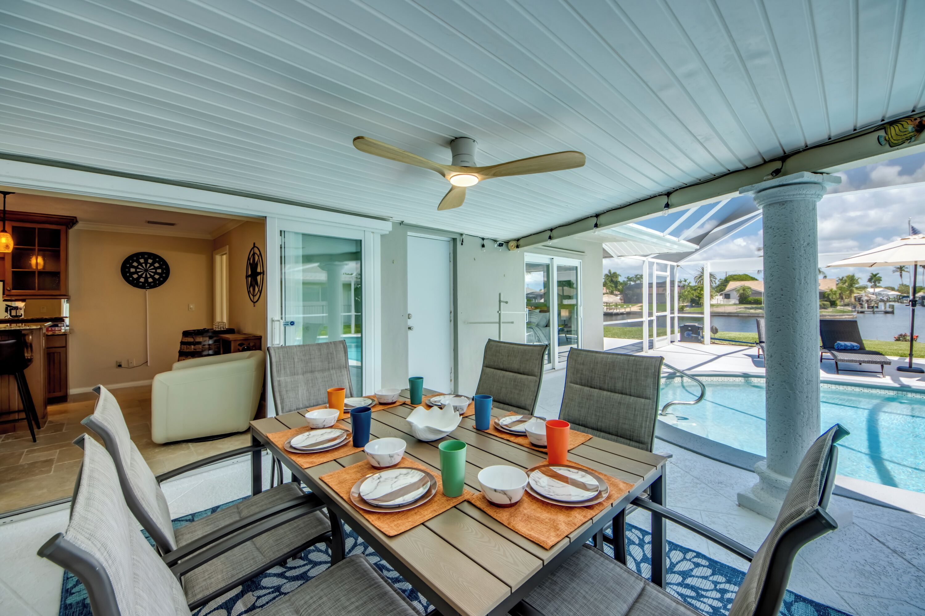 A covered outdoor dining area with a pool view, a ceiling fan, and a kitchen area with a dartboard.