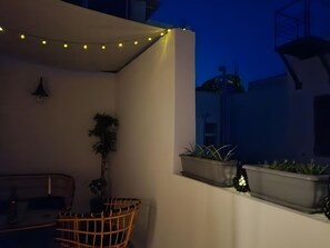 Ein Balkon mit Lichterketten, einem Korbstuhl, Topfpflanzen und einer wandmontierten Lampe.