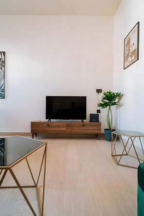 A modern living room with a flat-screen TV on a wooden stand, a glass coffee table, a potted plant, and a framed picture on the wall. - Casa Autentica 1933 Apartment with Private Balcony (Kos)