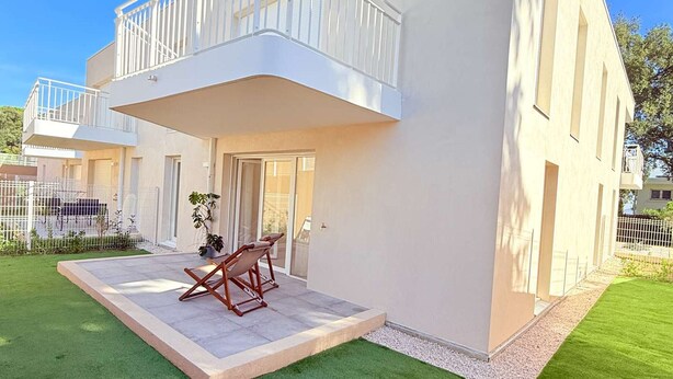 Un edificio residencial moderno de dos pisos, con un balcón, un pequeño patio con una silla y un césped bien cuidado.