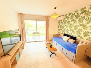 A furnished room with a sofa, a TV, a table with a plant, and a view through a sliding door to an outdoor area. - Apartment, air conditioning, parking - 4 guests (Le Lavandou)