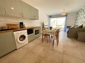 A compact kitchen with a washing machine, microwave, and coffee maker. A dining table with chairs is set up in the center. - Apartment, air conditioning, parking - 4 guests (Le Lavandou)