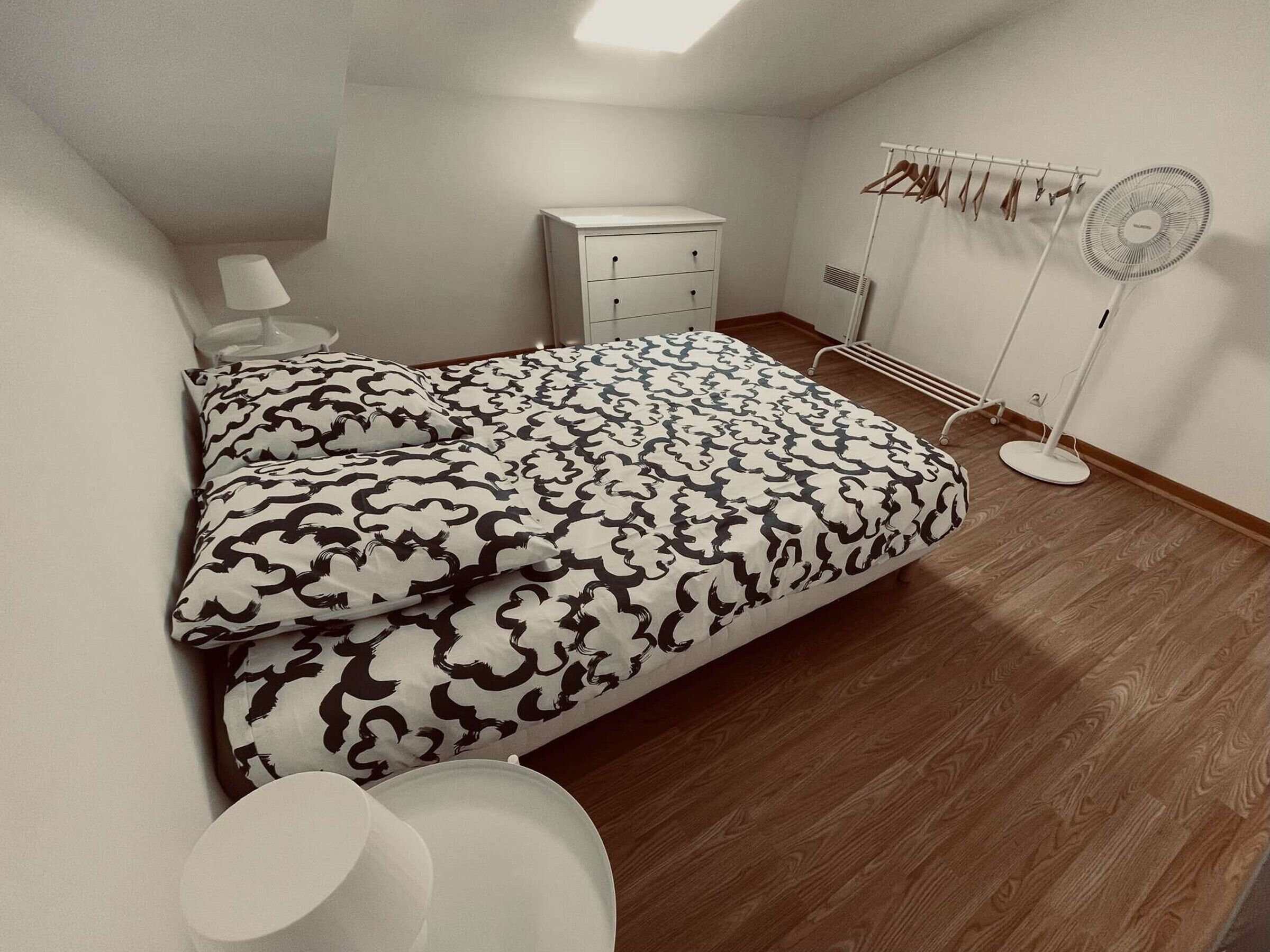 A bedroom with a bed, bedside table, lamp, wardrobe, and a standing fan.