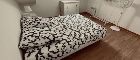 A bedroom with a bed, bedside table, lamp, wardrobe, and a standing fan.
