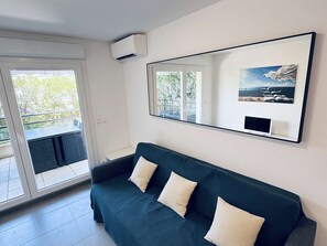 A modern living room with a dark green sofa, a large mirror, and a view of greenery outside. - T2 Apartment with pool and AC - spacious lodging (Le Lavandou)
