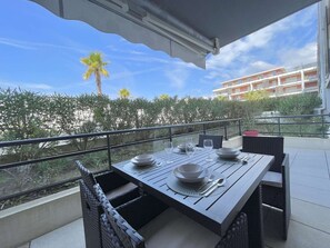 A dining area with a table set for two, outdoor furniture, and a view of a building and palm trees. - T2 Apartment with pool and AC - spacious lodging (Le Lavandou)