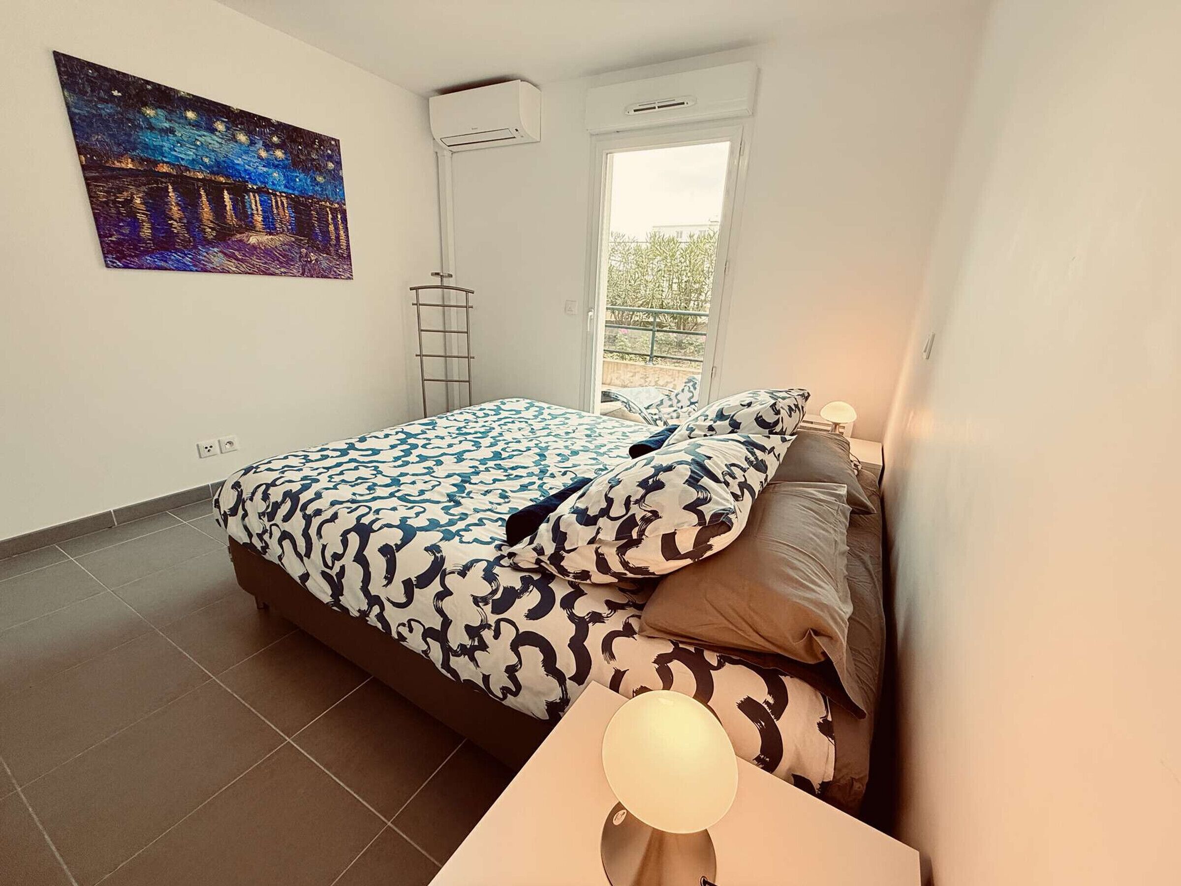 Ein Schlafzimmer mit einem Bett, einem Nachttisch mit Lampe, einem Fenster und einer Klimaanlage.