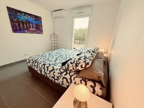 Une chambre à coucher comprenant un lit, une table de chevet avec une lampe, une fenêtre et un appareil de climatisation.