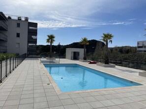 A swimming pool with a tiled deck, surrounded by a low fence, and palm trees in the background. - T2 Apartment with pool and AC - spacious lodging (Le Lavandou)
