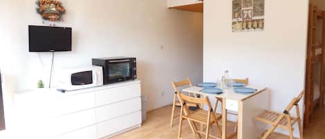 A compact living space with a dining area, a white cabinet with a microwave, a flat-screen TV, and a small plant.