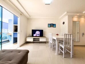 A modern living room with a flat-screen TV, a dining table with chairs, a sofa, and a large window offering a view of the outside. - Mont Blanc by LikeHome (Bombinhas)