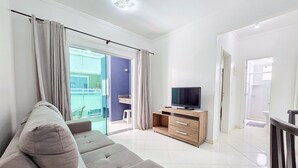 Ein modernes Wohnzimmer mit grauem Sofa, einem Flachbildfernseher auf einem Holzkabinett und einer Glasschiebetür, die auf einen Balkon führt.