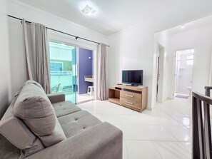 A modern living room with a grey sofa, a flat-screen TV on a wooden cabinet, and a sliding glass door leading to a balcony. - Mont Blanc by LikeHome (Bombinhas)