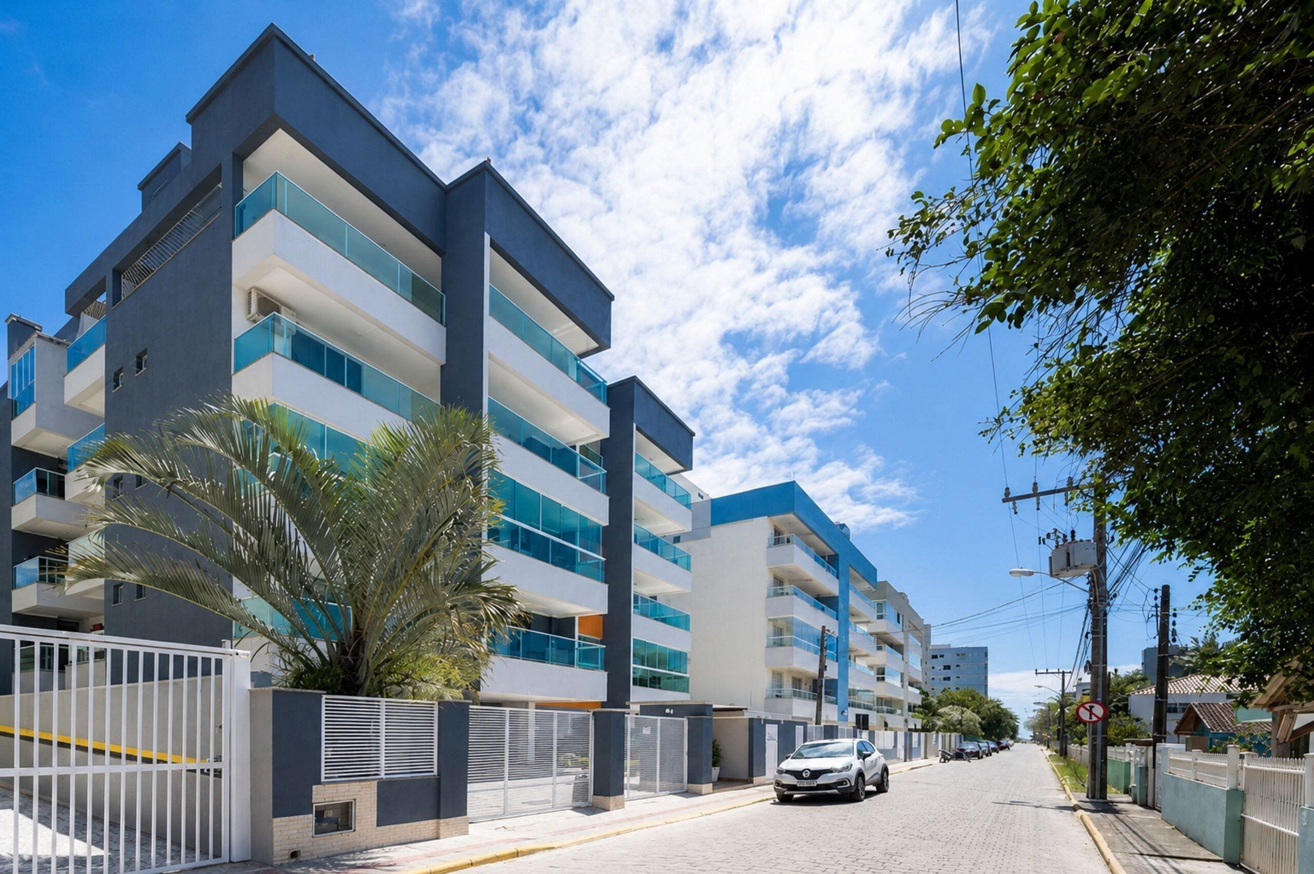 Um edifício residencial moderno, com várias andares, varandas, portão fechado e céu claro.