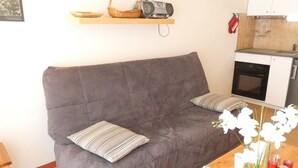 A grey sofa with striped pillows, a wooden table with a book and a potted plant, and a kitchen area with a microwave and oven. - Cozy apartment facing south - 4 guests (Saint-Lary-Soulan)