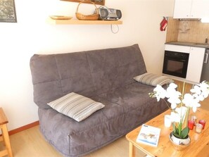 Ein graues Sofa mit gestreiften Kissen, ein Holztisch mit einem Buch und einer Topfpflanze, sowie eine Küchenzeile mit Mikrowelle und Backofen.
