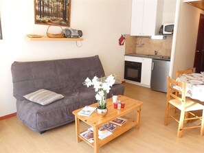 A living room with a grey sofa, a wooden coffee table with magazines, a small potted plant, and a kitchen area with a microwave and oven. - Cozy apartment facing south - 4 guests (Saint-Lary-Soulan)